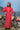Woman in a red dress standing next to a motorcycle by the ocean in lombok indonesia.  wearing a broderie anglaise kaftan in bright red cherry colour, and pure silk headband with custom artwork. 