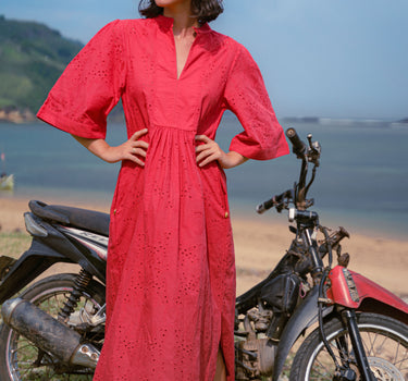 Woman in a red dress standing next to a motorcycle by the ocean in lombok indonesia.  wearing a broderie anglaise kaftan in bright red cherry colour, and pure silk headband with custom artwork. 